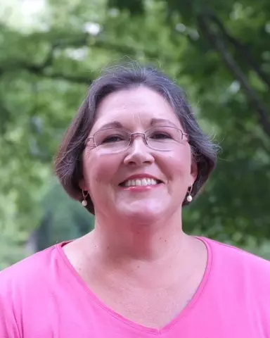 short brown hair, glasses, wearing a pink shirt; trees in the background